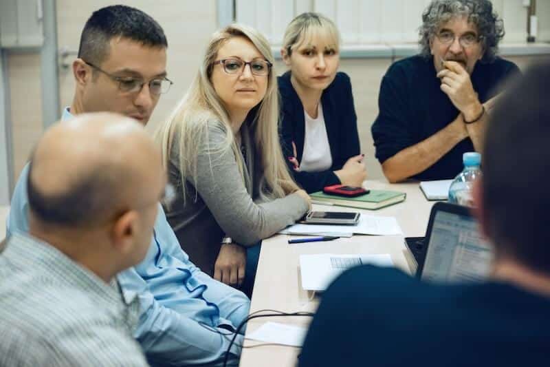 Six businesspeople sit around a table listening to one of their number, representing how according to subsidiarity people should take personal responsibility but work together