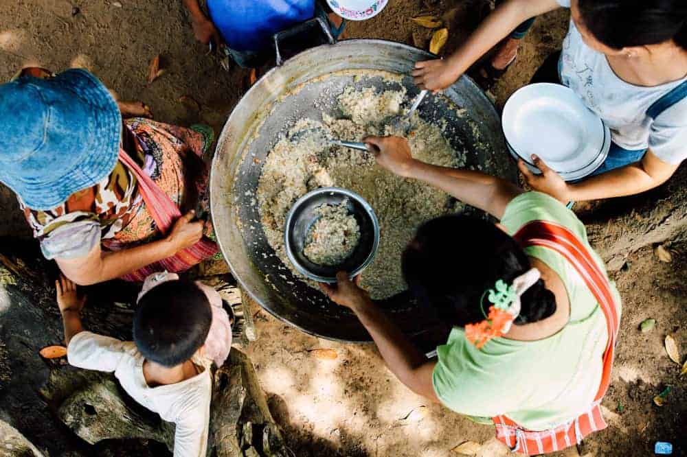 A family stands around a common dish of food above a dirt floor, showing how according to the universal destination of goods we are all equally responsible for each other