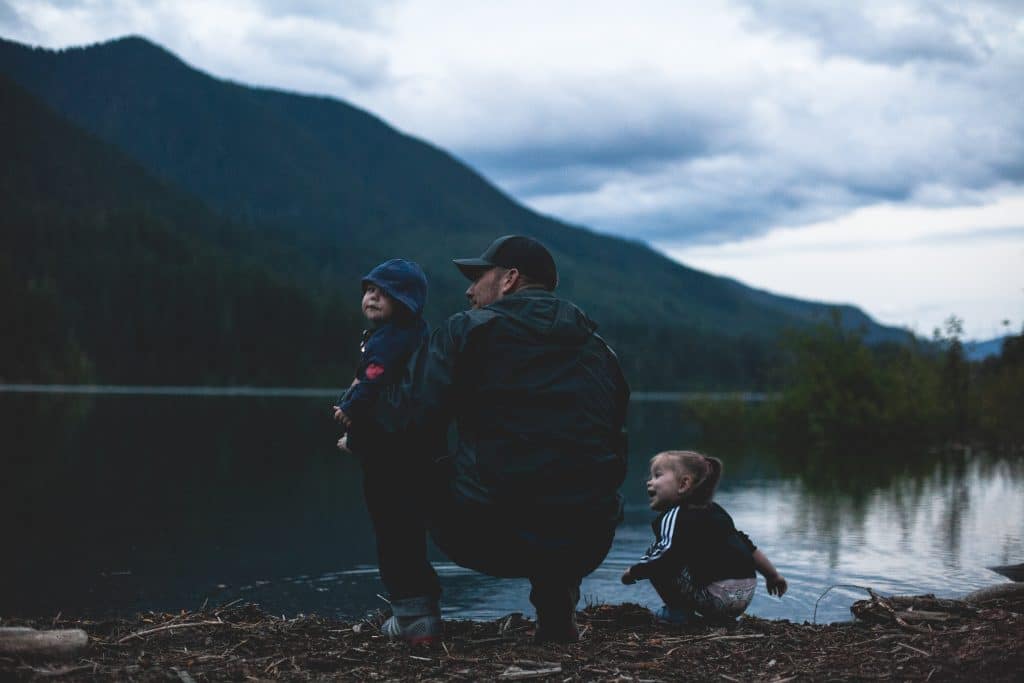 A man with two kids plays near a large lake with forested mountains on the other side, showing the unity of the human and physical environment