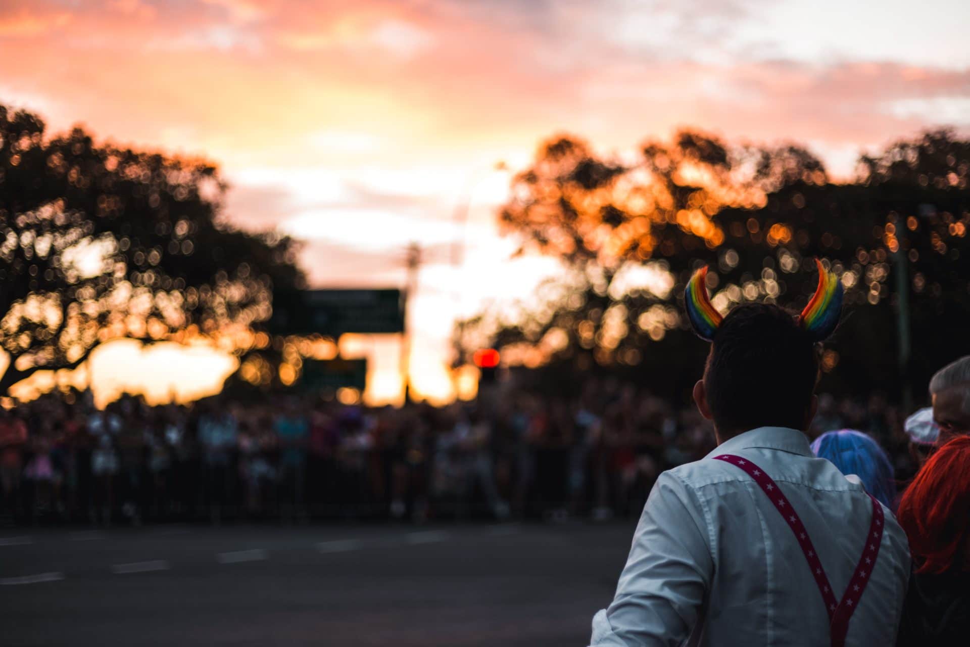 A man with a white shirt and rainbow demon horns stands with his back toward the viewer at a protest in the evening, showing the chaos that results from destroying the human environment