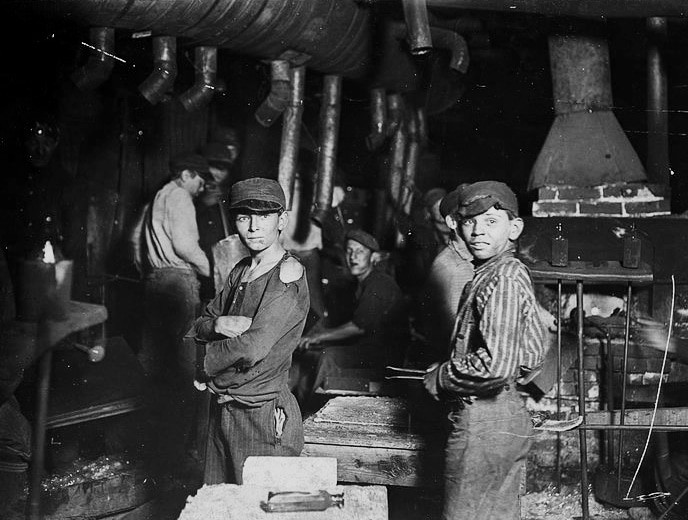 What is Catholic social teaching? in a black and white photograph two boys in poor clothes stand in a glassworks
