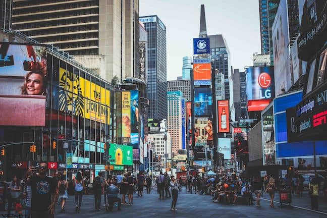 People walk along a city street surrounded by billboards, showing the effect of consumerism on society as a whole